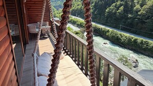A wooden deck with a rope swing, a cushioned bench, and a view of a river and forest. - Hira Bungalov (Camlihemsin)