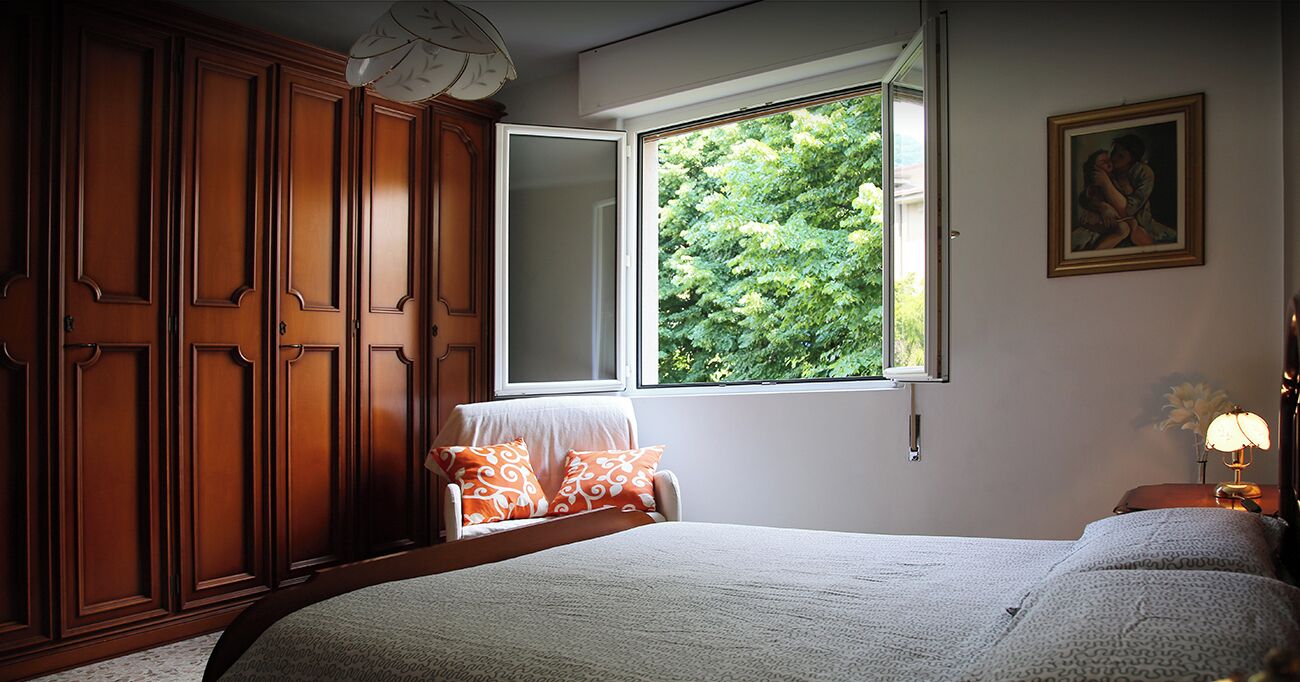 A bedroom with a large bed, a window with a view of greenery, a wardrobe, and a bedside table with a lamp.