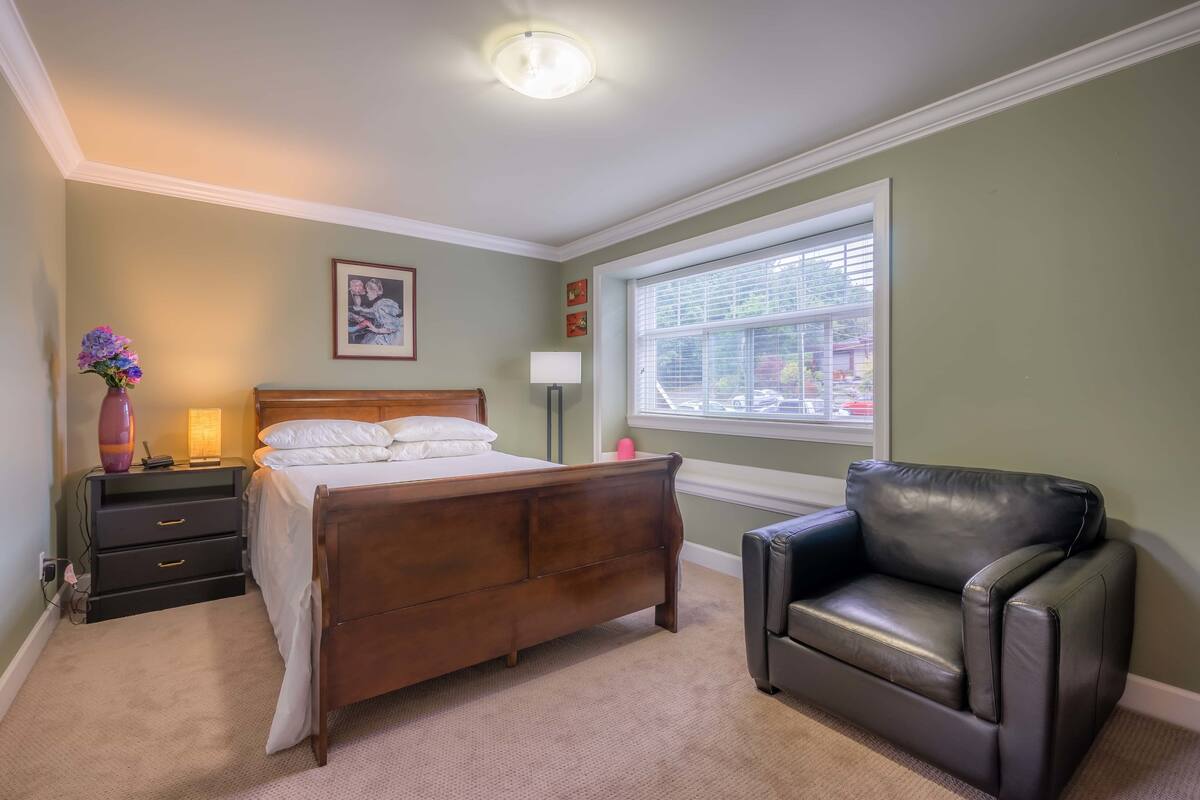Een slaapkamer met een houten bed, een zwarte leren fauteuil, een nachtkastje met lades, een bijzettafeltje met lamp en een raam met jaloezieën.