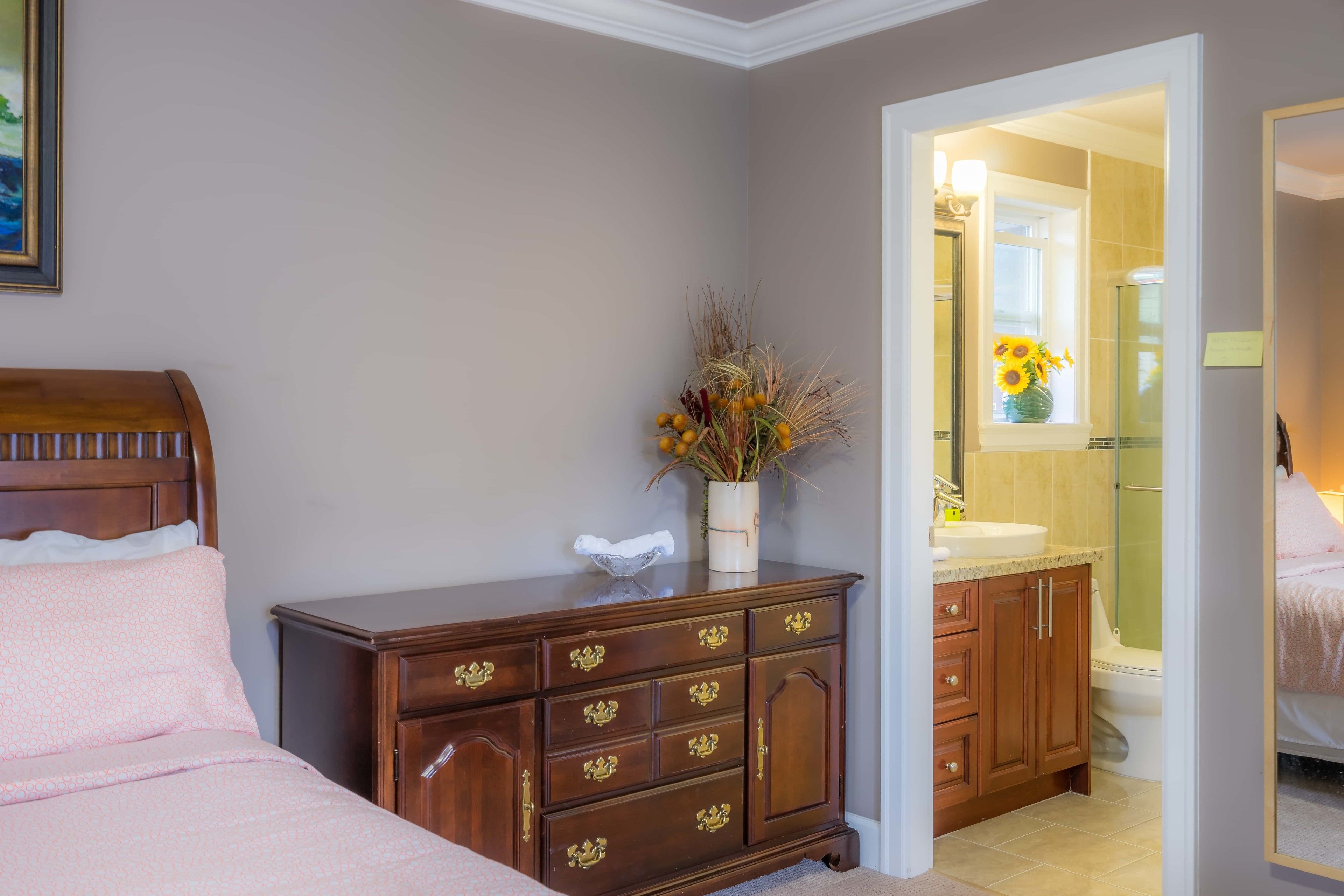 Une chambre avec une commode en bois, un lit avec une literie rose et une salle de bain visible à travers une porte ouverte.