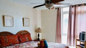 A bedroom with a bed, ceiling fan, lamp, TV stand, and curtains. - Special December rates /Spectacular Ocean views 1Bed/2 bath (Kihei)
