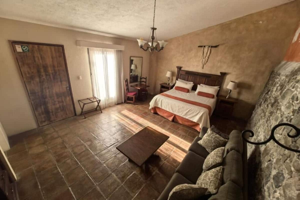 A bedroom with a bed, a sofa, a wooden coffee table, a chandelier, and a stone wall.