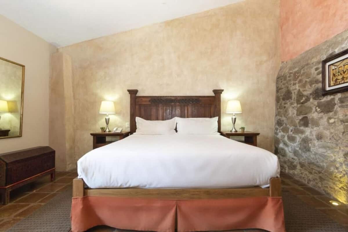 A single bed with a wooden headboard, two bedside lamps, a nightstand, and a stone wall.