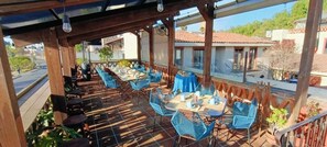 An outdoor patio with tables and chairs, a covered area with a view of the street, and a building in the background.
