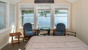 Quarto com uma cama grande, duas cadeiras azuis, uma mesinha e vista para um lago através de três janelas.