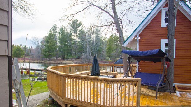 Terraza de madera con una silla mecedora y un área de estar techada, con vista a un cuerpo de agua y un área boscosa.