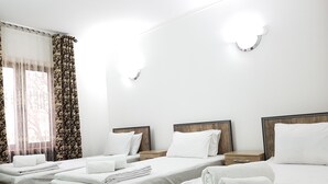 Une chambre d’hôtel avec deux lits simples et un lit plus grand, tous dotés de draps et d’oreillers blancs. On y trouve des têtes de lit en bois, une petite table de chevet en bois et un tapis à motifs verts sur le sol.