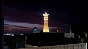 Une vue nocturne d’une ville, avec une tour illuminée qui se détache sur le paysage et un bâtiment orné de sculptures architecturales complexes.