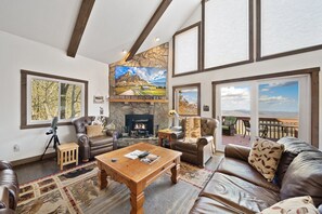 A spacious living room with leather sofas, a fireplace, a stone accent wall, and large windows offering views of the outdoors. - High Country Escape with Views (Seven Devils)