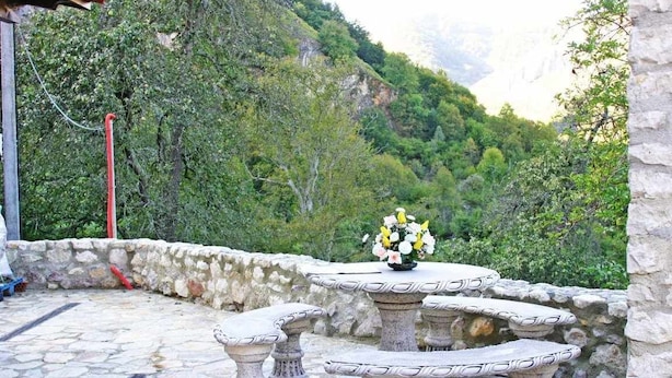 A stone patio with a table and benches, surrounded by a stone wall and lush greenery.