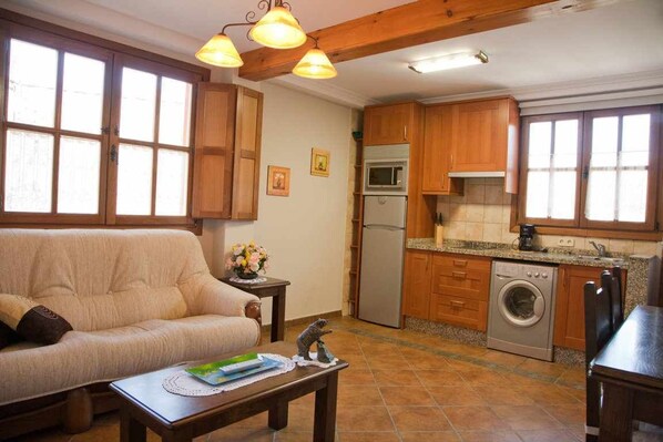 Una sala de estar acogedora con sofá, mesa de centro y una zona de cocina que cuenta con armarios de madera, lavadora y refrigerador.