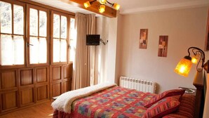 A bedroom with a wooden floor, a bed with a colorful quilt, a white radiator, a TV mounted on the wall, and a window with curtains.