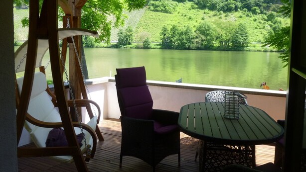 A wooden deck with a swing, two chairs, and a table overlooking a lake and forest.