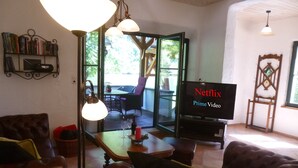 A cozy living room with a leather couch, a coffee table, and a TV displaying 'Netflix Prime Video'. There is a bookshelf, a window with a view of greenery, and a wooden chair.