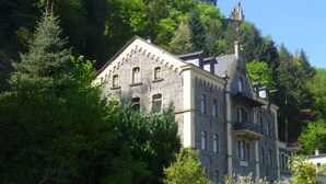A historic stone building with multiple windows and a turret, surrounded by dense greenery and trees.
