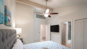 Un dormitorio con cabecera capitonada, edredón estampado, ventilador de techo, televisor de pantalla plana y una puerta que da a otra habitación.