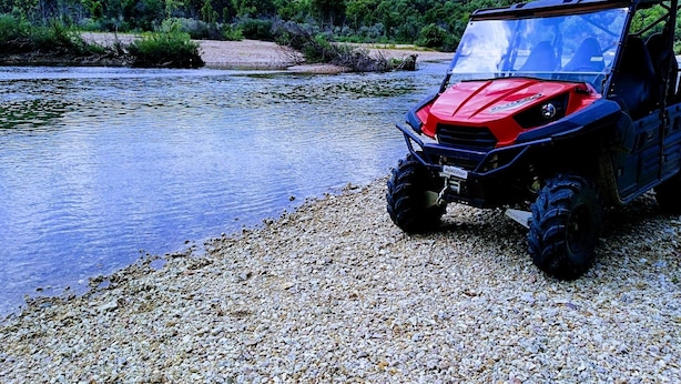 Un vehículo todo terreno rojo estacionado en un camino de grava junto a un río, con un paisaje boscoso al fondo.