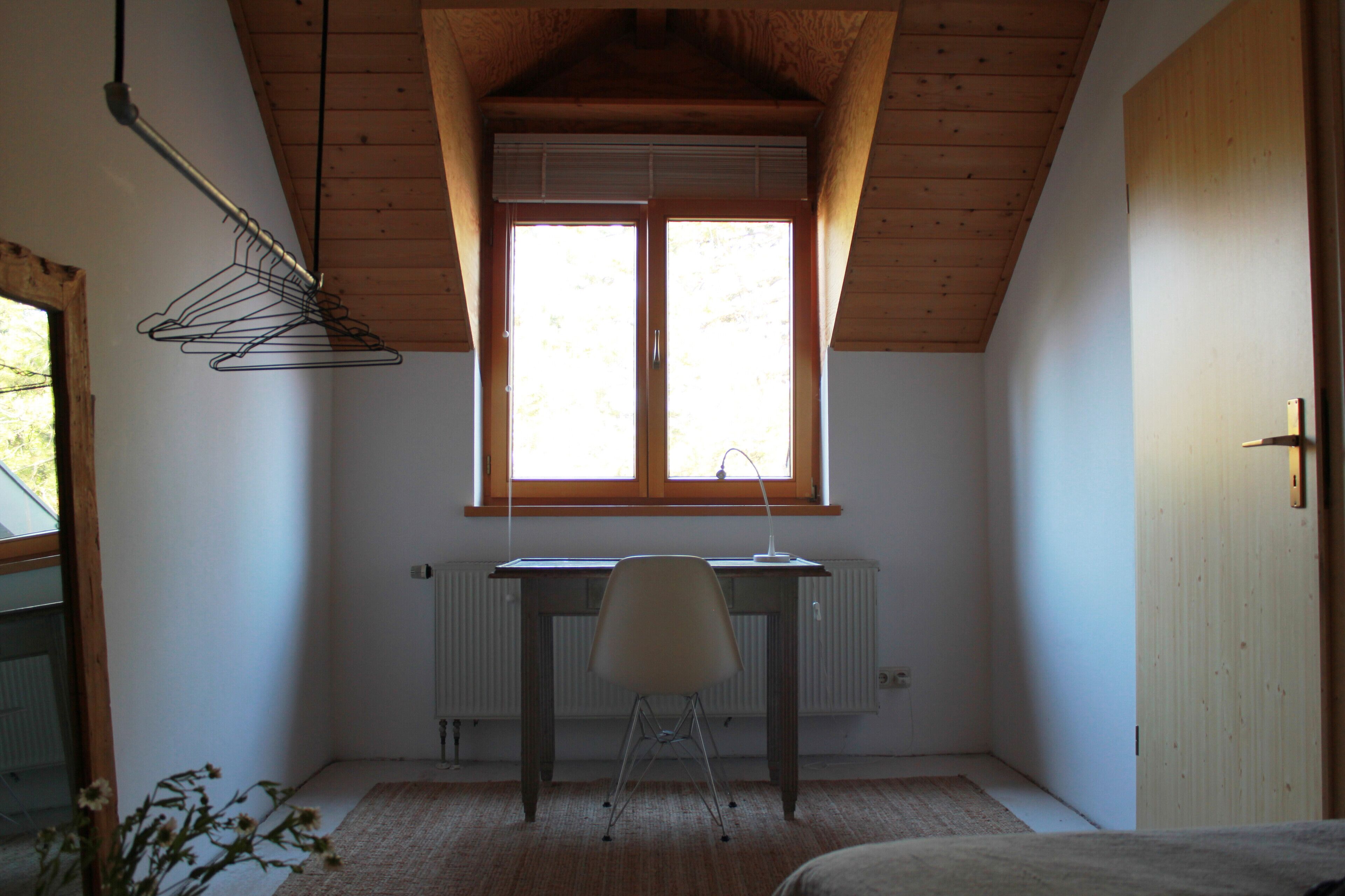 Une pièce avec un plafond en pente, un radiateur, un bureau avec une chaise et une fenêtre avec des stores.