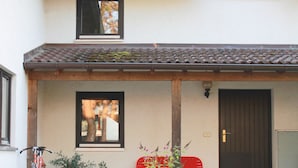 Ett tvåvåningshus med ett tak täckt av tegelpannor, en ytterdörr och en liten veranda med en röd stol och krukväxter.