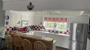 Una cocina con armarios blancos, un refrigerador de acero inoxidable, una isla de madera con taburetes de bar, una ventana con un visillo rojo y blanco a cuadros, y un reloj montado en la pared.