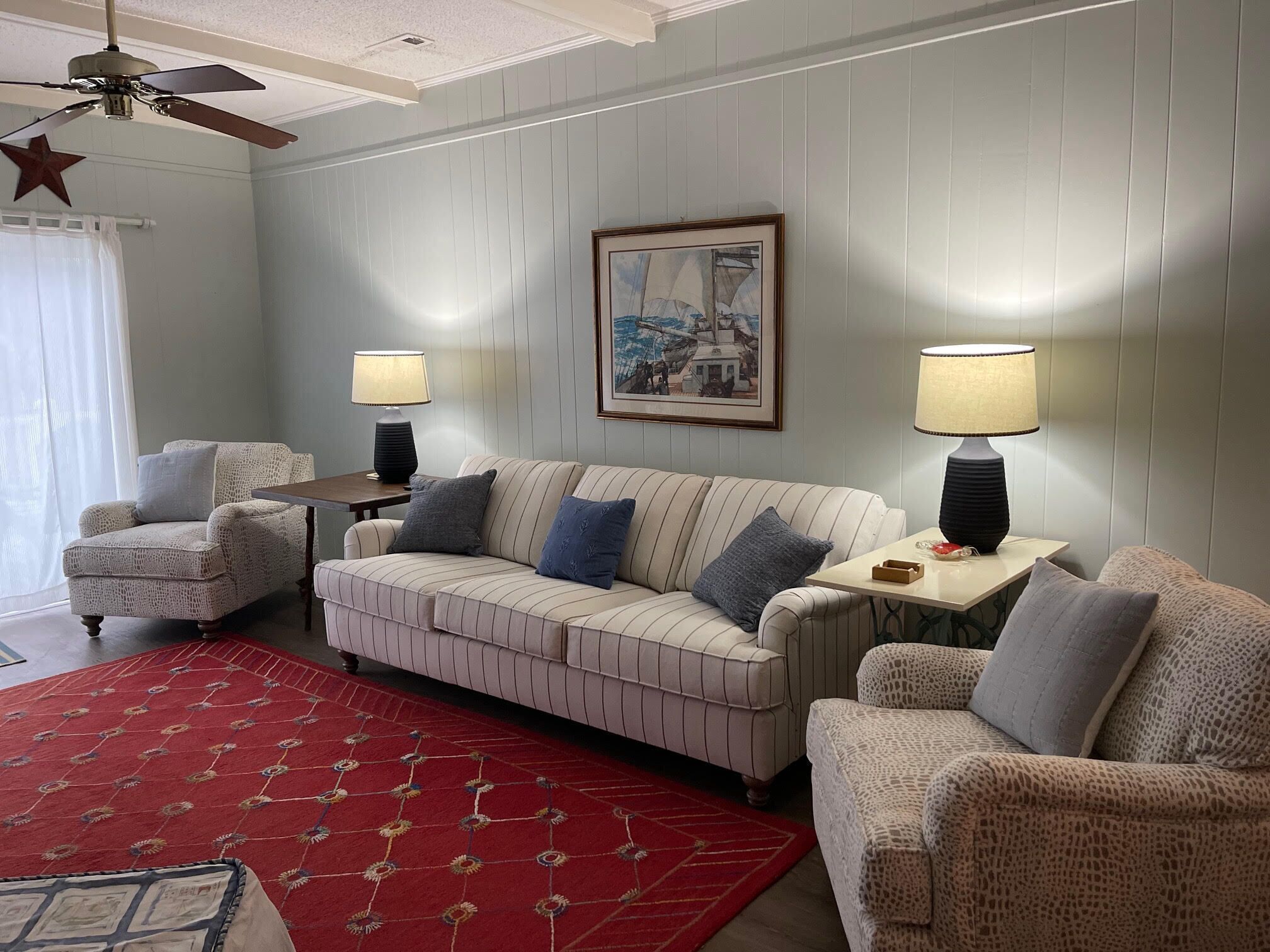 A living room with a striped sofa, two armchairs, a red patterned rug, a ceiling fan, and two table lamps.