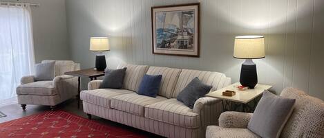 A living room with a striped sofa, two armchairs, a red patterned rug, a ceiling fan, and two table lamps.