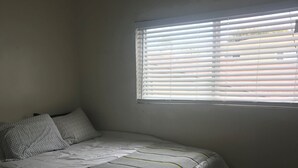 Une petite pièce peu éclairée, avec un lit simple, une fenêtre munie de stores et un mur uni.