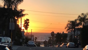 Straatbeeld bij zonsondergang met uitzicht op de oceaan, palmbomen en geparkeerde auto's.
