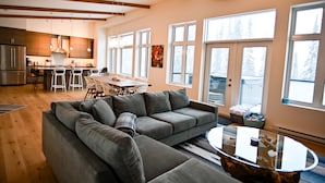 A modern living room with a sectional sofa, a glass coffee table, and a dining area with a wooden table and chairs. - New built, modern ski-in/ski-out home with great views of Columbia River Valley (Golden)