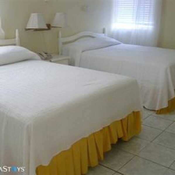 Habitación de hotel con dos camas individuales, una mesita de noche con lámpara y una ventana con persianas.