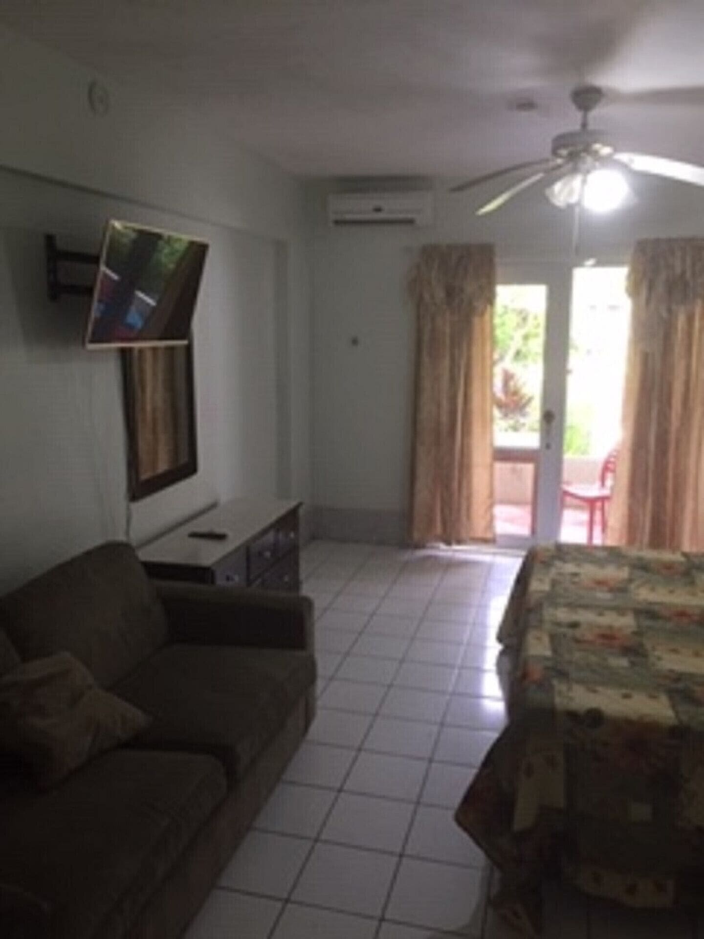 Una sala de estar con un sofá, un televisor de pantalla plana montado en la pared, un ventilador de techo y vistas al exterior a través de un ventanal.