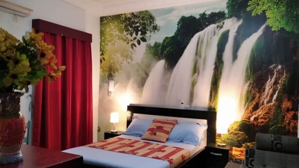 Ein Hotelzimmer mit Bett, roten Vorhängen, einem Wandbild mit einem Wasserfall und einem Nachttisch mit Lampe.
