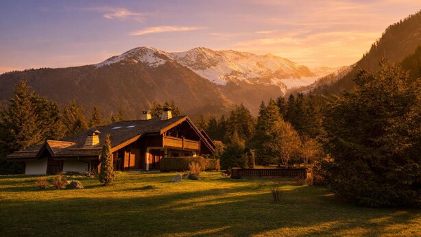 Une cabane dotée d’un toit en pente, d’une façade en bois et d’une cheminée, entourée d’une zone herbeuse et d’arbres, avec des montagnes en arrière-plan.