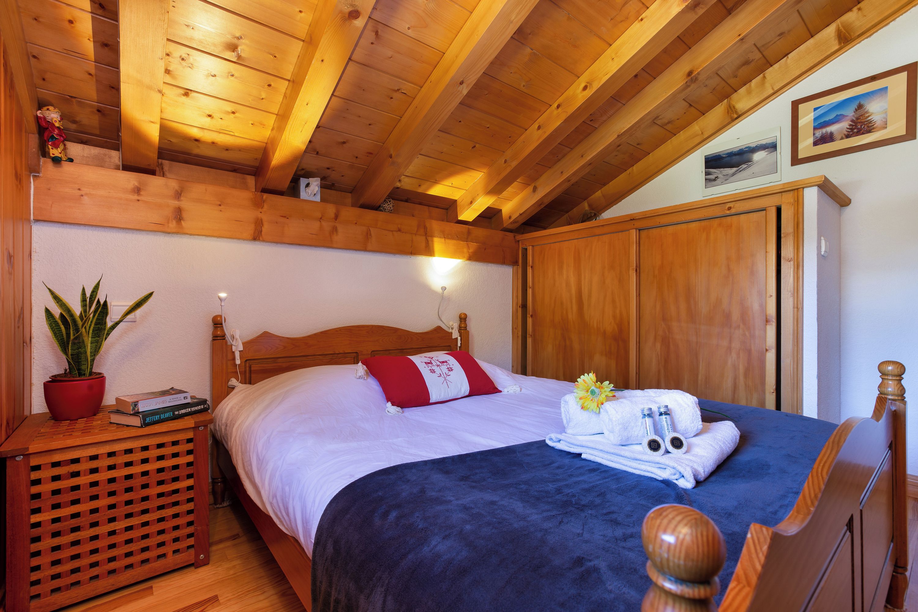 A wooden-paneled room with a bed, a nightstand, a potted plant, and a wall-mounted light.