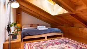 Une chambre mansardée chaleureuse avec un plafond en pente, un lit recouvert d’une couverture grise, une plante en pot et un tapis coloré.