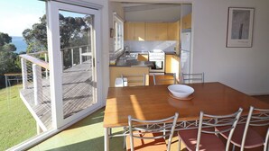 A modern kitchen with a view of the sea, a dining area with a wooden table and metal chairs, and a sliding glass door leading to a deck. - 60 Karbeethong Avenue, Mallacoota (Mallacoota)