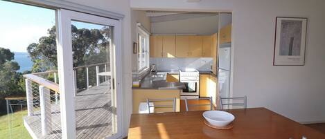 A modern kitchen with a view of the sea, a dining area with a wooden table and metal chairs, and a sliding glass door leading to a deck.