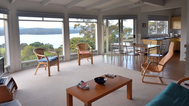 A spacious living area with a large window overlooking a lake, furnished with wicker chairs and a wooden table. - 60 Karbeethong Avenue, Mallacoota (Mallacoota)