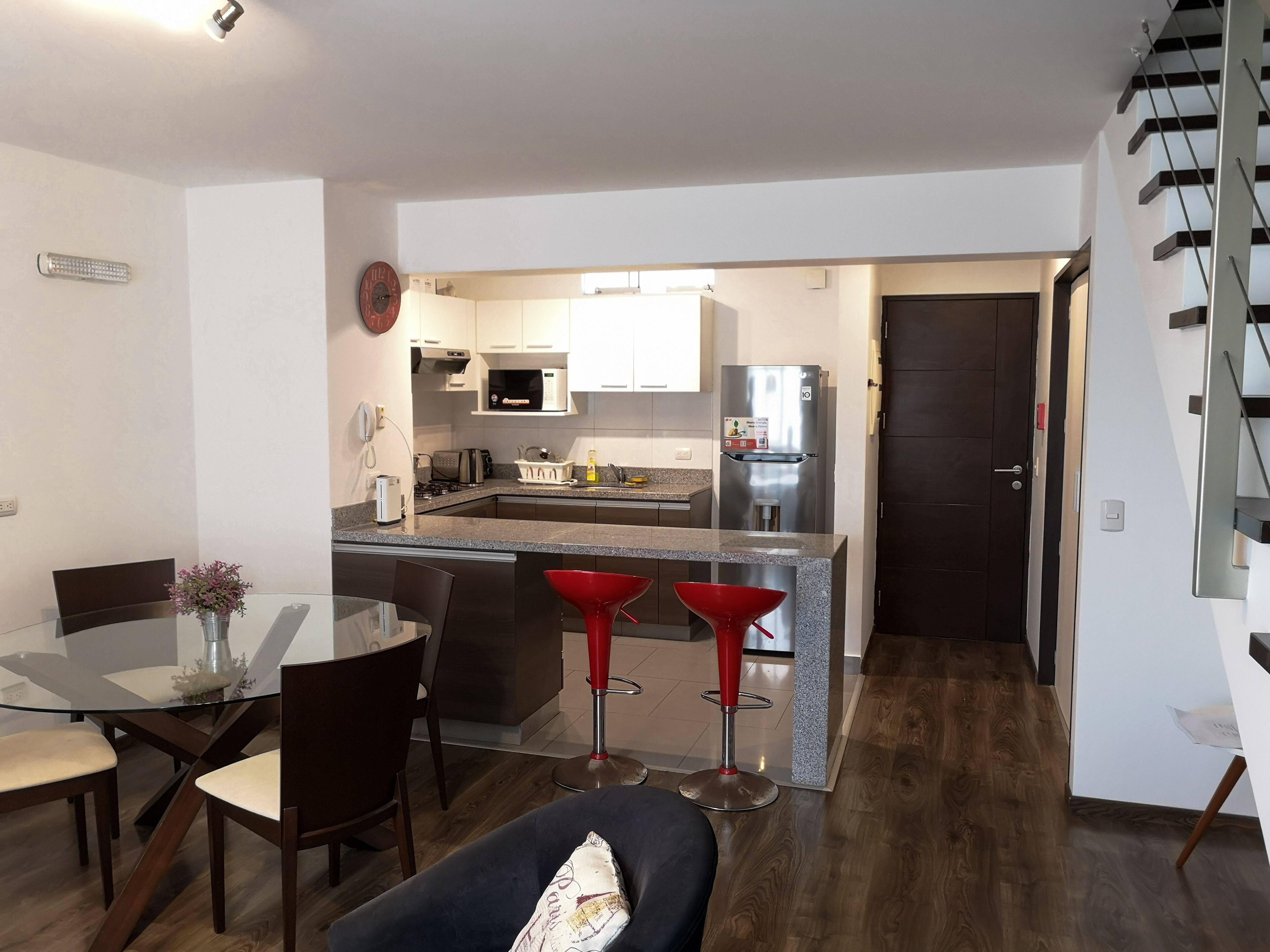 A modern kitchen with a central island, a dining area with a glass table and chairs, and a staircase to the right.