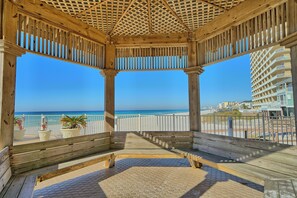 Relax in a serene beachside gazebo with a clear ocean view, surrounded by lush greenery and a white fence.