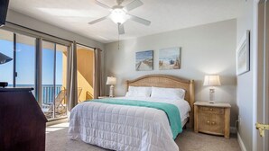 A bedroom with a bed, two bedside lamps, a ceiling fan, and a view of the ocean through large windows.