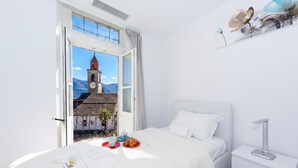 A hotel room with a bed, a bedside table, a window with a view of a building and mountains, and a breakfast tray on the bed. - Yellow view apartment - Ronco S/Ascona, Ronco sopra Ascona, Switzerland (Ronco sopra Ascona)