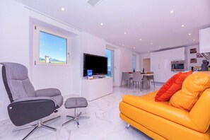 A modern living room with a grey office chair, an orange sofa, a flat-screen TV, and a dining area in the background. - Yellow view apartment - Ronco Sopra Ascon, Ronco sopra Ascona, Switzerland (Ronco sopra Ascona)