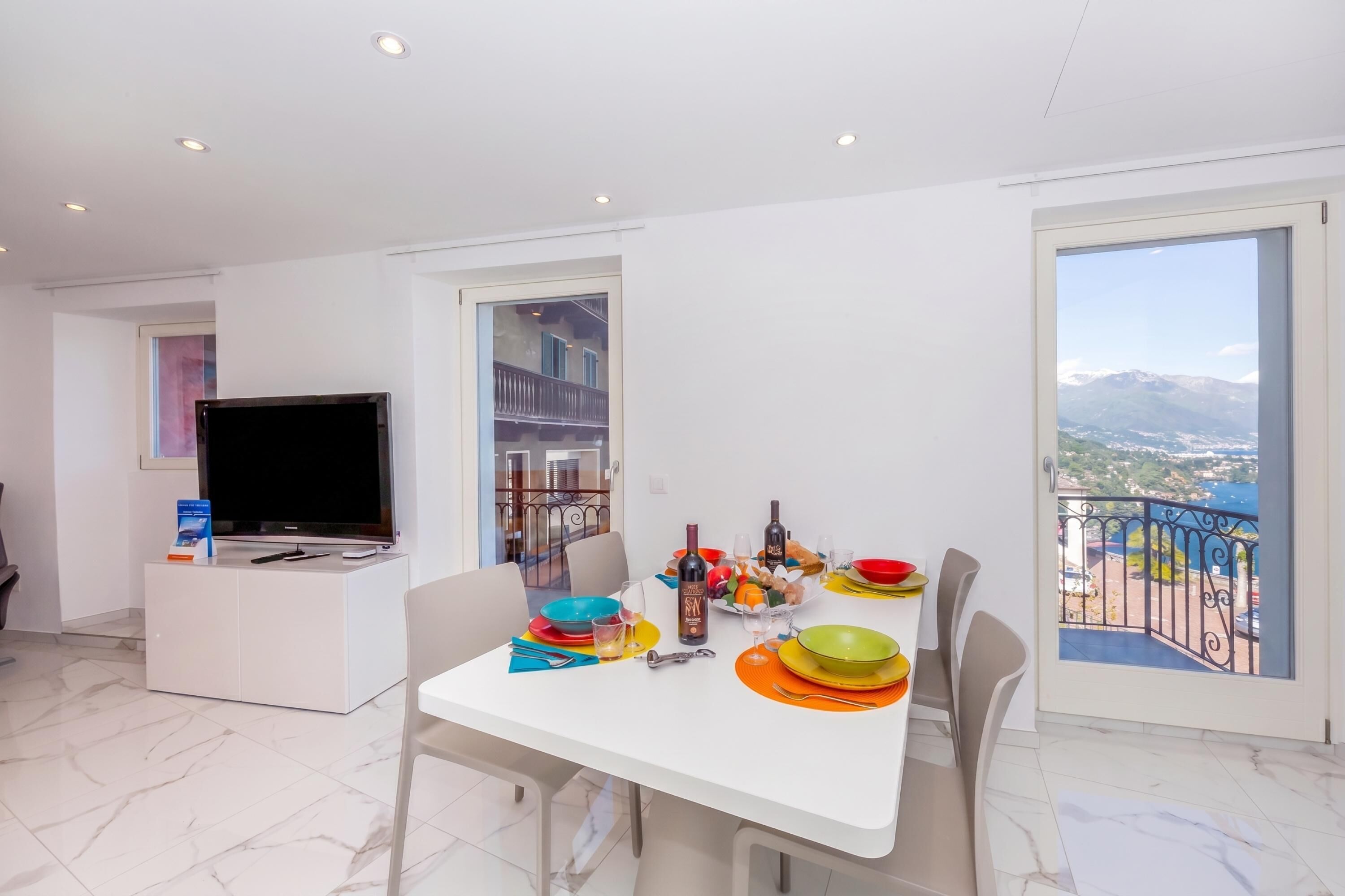 A modern dining area with a TV, dining table, chairs, and a balcony with a mountain view.