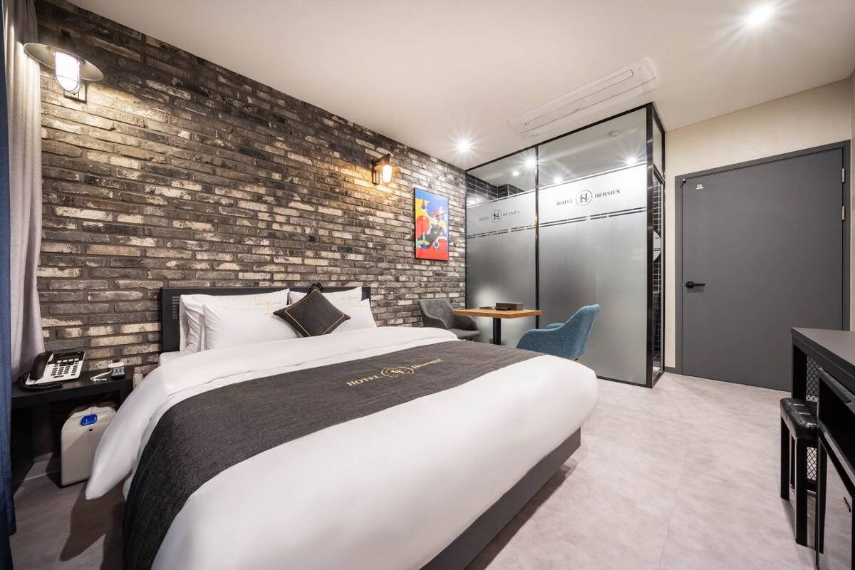 A modern hotel room with a brick-patterned wall, a bed with white bedding, a desk with a chair, and a door labeled "HOTEL HERMES".