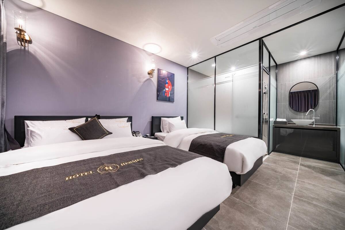 A hotel room with two beds, a glass-enclosed shower area, and a small bathroom with a sink and mirror.