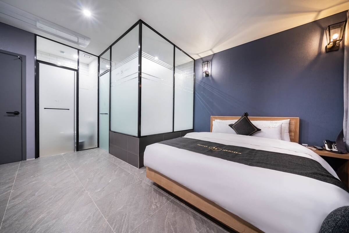 A modern hotel room with a large bed, a bedside table with a lamp, and a glass partition wall.