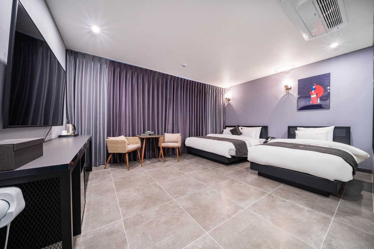 A hotel room with two beds, a desk, a chair, a small table, and a wall-mounted TV.