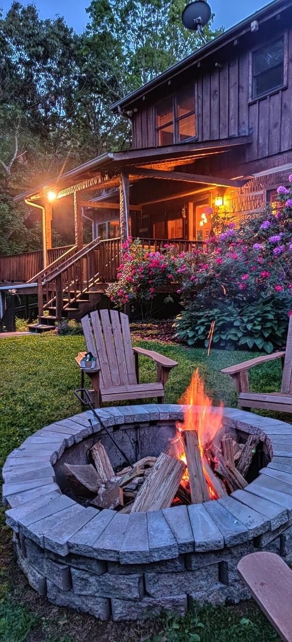 A wooden cabin with a stone fire pit in front, surrounded by lush greenery and flowering plants.