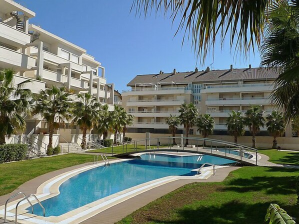 A swimming pool with a curved design, surrounded by palm trees and a multi-story building with balconies.